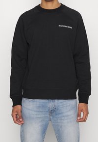 Sudadera negra con cuello redondo y manga larga. Presenta puños y dobladillo de canalé, y un logo blanco en el pecho. Textura de tela suave.