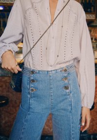 Lys hørskjorte med lodrette udskæringer og knapper parret med højtaljede blå denimjeans med guldfarvede knapdetaljer.