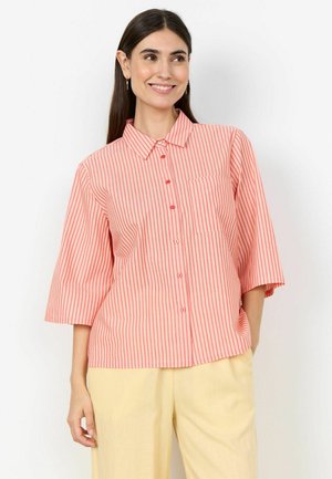 Smiling woman with long dark hair wearing pink and white vertical striped button-up shirt with elbow-length sleeves and light yellow pants.
