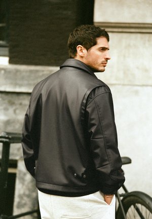 Mężczyzna w czarnej skórzanej kurtce i jasnych spodniach stoi na zewnątrz obok roweru, patrząc na bok z rękami w kieszeniach.