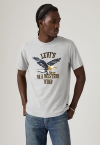 Jonge man met gevlochten haar, gekleed in een grijze Levi's T-shirt met een vliegende adelaar en de tekst "On a Western Wind", gecombineerd met blauwe jeans.