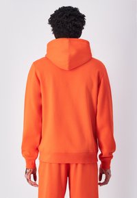Sudadera naranja hecha de tejido suave, con capucha ajustable y puños acanalados. La parte trasera es lisa con un ajuste ligeramente relajado.