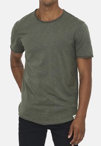 Olivgrünes T-Shirt mit kurzen Ärmeln und rundem Ausschnitt, ausgestattet mit einer subtilen Textur und kontrastierenden Nähten am Saum und an den Ärmeln.