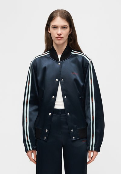 Γυναίκα που φοράει μπλε σατέν bomber μπουφάν με λευκές ρίγες και κόκκινο λογότυπο στα μανίκια, συνδυασμένο με παντελόνι σε αντίστοιχο μπλε και λευκό τοπ.