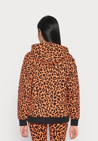 Oranje hoodie met zwarte luipaardprint, lange mouwen en een capuchon met trekkoord. Heeft een zwarte zoom en manchetten, gecombineerd met bijpassende leggings.