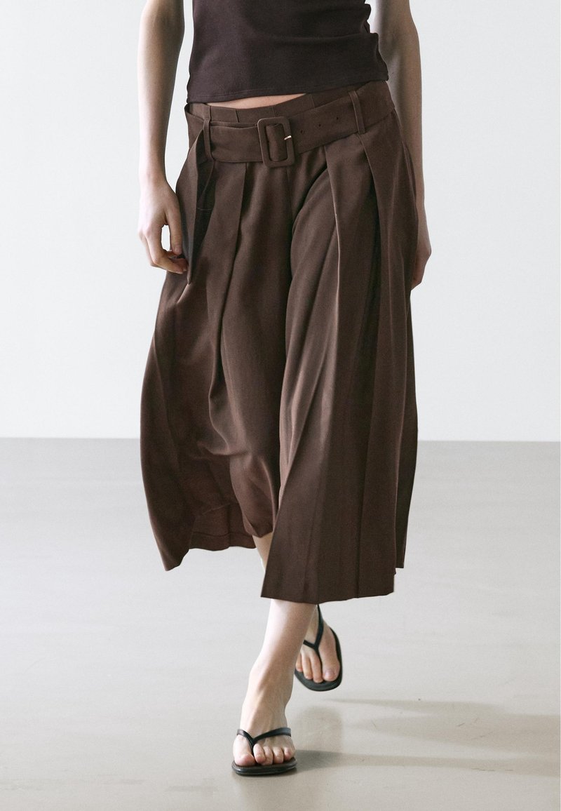 Personne portant un pantalon large marron raccourci avec une ceinture assortie et des sandales noires à bride, marchant sur un sol lisse.