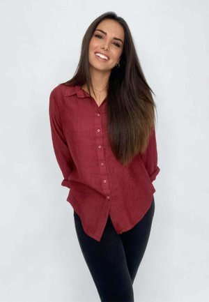 Giovane donna con lunghi capelli scuri sorridente, che indossa una camicia bordeaux con bottoni e pantaloni neri, in piedi davanti a uno sfondo chiaro e semplice.