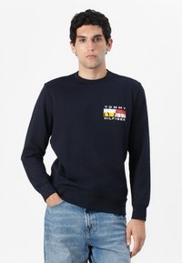 Mørkeblå sweatshirt laget av bomullsblanding, med rund hals og ribbestrikkede mansjetter. Har en fargerik logo på venstre brystområde.