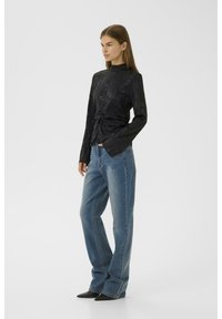 Haut noir à manches longues avec un motif floral texturé et une taille nouée, associé à un jean droit bleu clair et des chaussures noires à bouts pointus.