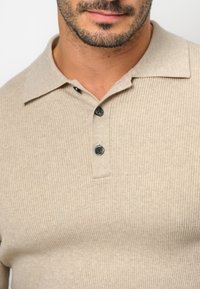 Beige Ripp-Polohemd mit einem lockeren Kragen und drei dunkelgrünen Knopfverschluss am Ausschnitt. Weiche Textur und taillierte Passform.