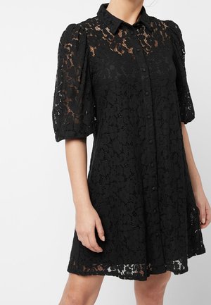 Krótka czarna sukienka z koronki w kwiatowy wzór, z przodu zapinana na guziki i z bufkami na rękawach, prezentowana na stojącej osobie na jednolitym tle.