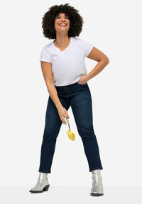 Bílý krátký rukáv tričko, modré skinny džíny a lesklé stříbrné kotníkové boty. Model drží žlutý předmět, usmívá se a má kudrnaté vlasy.