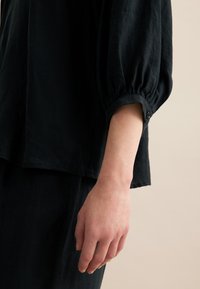 Top nero con texture e vestibilità ampia. Presenta maniche a sbuffo e un dettaglio con bottone al polsino. Design semplice e minimalista.