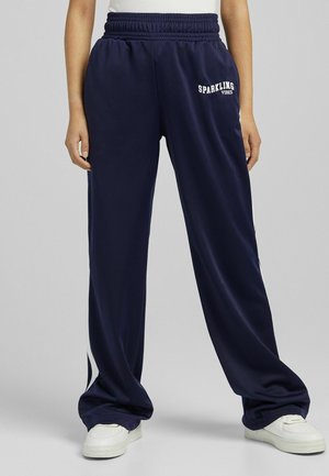 Osoba nosząca granatowe szerokie spodnie z elastycznym pasem i białym napisem „SPARKLING VIBES”, w połączeniu z białymi trampkami.