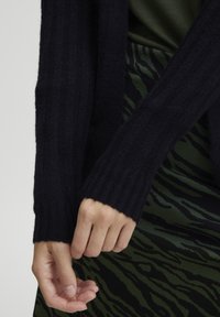 Mörk marinblå ribbad stickad cardigan med långa ärmar, med ett strukturerat mönster. Händerna är lätt placerade mot en grön zebra-tryckt kjol.