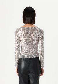 Μακρυμάνικο τοπ σε ασημί στρας με στενή σιλουέτα. Η πλάτη διαθέτει στρογγυλή λαιμόκοψη και διακριτή ραφή, συνδυασμένο με μαύρο παντελόνι.