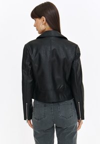 Czarna kurtka z syntetycznej skóry o dopasowanym kroju, z wystającymi klapami i zamkami na mankietach. Gładka tekstura i skrócona długość, w zestawieniu z szarymi dżinsami.