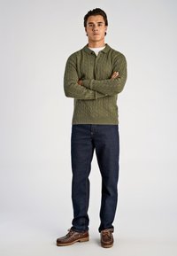 Olivengroene gebreide polotrui met een gestructureerd kabelpatroon, gecombineerd met donkerblauwe jeans en bruine bootsschoenen.