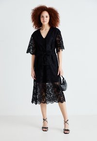 Robe en dentelle noire avec décolleté en V, manches courtes et ceinture à la taille. Présente un bas transparent avec des motifs complexes. Modèle tenant un petit sac.