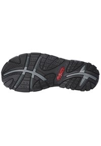 Semelle de chaussure en caoutchouc noir avec des crampons profonds, des accents gris et des motifs texturés. Un détail de logo rouge est visible près du centre.