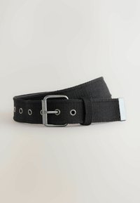 Ceinture en tissu noir tissé avec boucle en métal et plusieurs œillets en métal, posée enroulée sur un fond clair uni.