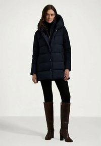 Γυναίκα με μαύρο παλτό τύπου puffer έως το γόνατο, μαύρο παντελόνι και καφέ μπότες μέχρι το γόνατο, να στέκεται σε απλό φόντο.