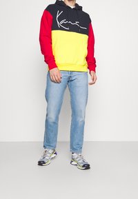 Felpa con cappuccio a blocchi di colore con pannelli blu navy, rossi e gialli, tasca anteriore e coulisse. Indossata con jeans azzurri e sneaker multicolore.