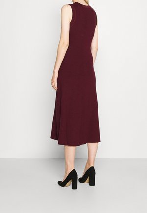 Kobieta w bezrękawnikowej, sięgającej kolan bordowej sukience i czarnych szpilkach, stojąca na tle jednolitego białego tła.