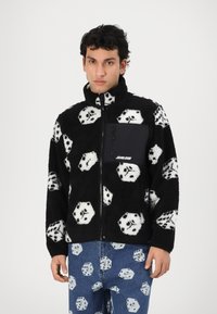 Black fleece jacket featuring white dice pattern, high collar, full zipper, and a solid black chest pocket with a logo. Denim pants with a dice pattern.