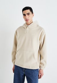 Beige fleece hettegenser med frontlomme, snor i hetten og elastiske mansjetter. Har en subtil logo på brystet. Brukes sammen med blå jeans.