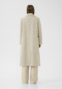 Manteau en laine beige avec un design long et droit, poches latérales et col cranté. Présente une fente à l'arrière, montrée de dos.