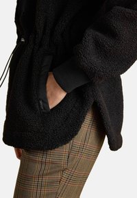 Ręka w kieszeni czarnej, teksturowanej kurtki polarowej z elastycznym mankietem, noszonej na brązowych spodniach w kratę.
