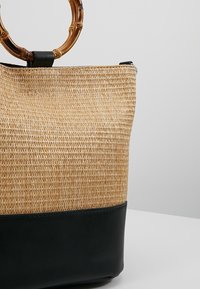 Borsa a tracolla in paglia con manico in bambù, caratterizzata da una parte superiore in fibra naturale testurizzata e una base in pelle nera solida. Chiusura superiore con cerniera.
