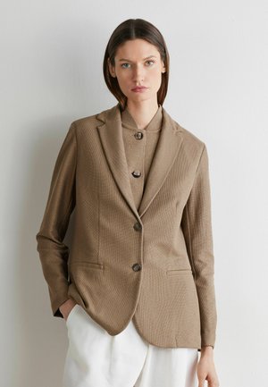 Frau mit glatten braunen Haaren, die ein beigefarbenes Blazer über einem geknöpften Oberteil und weißen Hosen trägt, steht vor einer schlichten, hellen Wand.