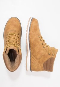 Botas de tobillo de ante marrón con cordones dorados, cuello acolchado y detalles texturizados, suela de goma y costuras beis.