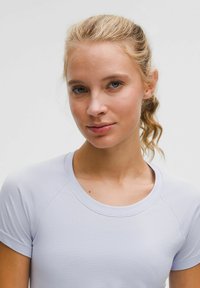 Jonge vrouw met blond haar in een paardenstaart, gekleed in een lichtpaarse t-shirt met korte mouwen, kijkt iets naar de zijkant tegen een effen achtergrond.