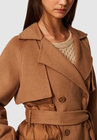 Dvojradový kabát v teplej hnedej vlnenej zmesi s odhaleným golierom, opaskom v oblasti pása a prešívanou hnedou podšívkou. Má veľké hnedé gombíky.