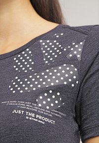 Camiseta gris oscuro con parches de tela de lunares en contraste, que presenta un diseño texturizado y texto impreso en la parte inferior.