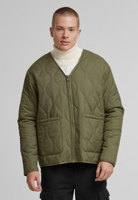 Olivgrüne gesteppte Jacke mit Reißverschluss vorne, rundem Kragen und zwei vorderen Taschen. Besitzt eine glatte Textur und eine Wellenmuster-Steppung.