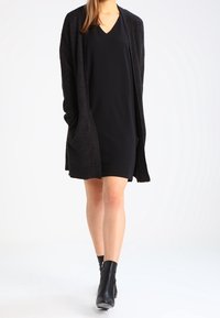 Mustat kardigans, joissa on teksturoitu pinta, mustan V-pääntiellä olevan mekon päällä. Asusteessa on nilkkapituiset mustat saappaat, luoden monochromaatista ilmettä.