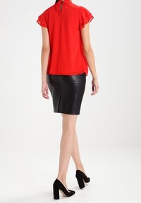 Czerwona bluzka z krótkim rękawem, z falbanami i wiązaniem przy szyi, w połączeniu z dopasowaną czarną skórzaną spódnicą. Czarne buty na wysokim obcasie.