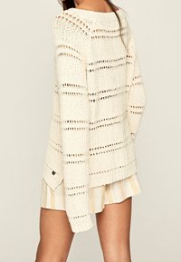 Cream, knitted sweater with open weave design, featuring horizontal stripes and long sleeves. Accented with a small black logo on the hem.