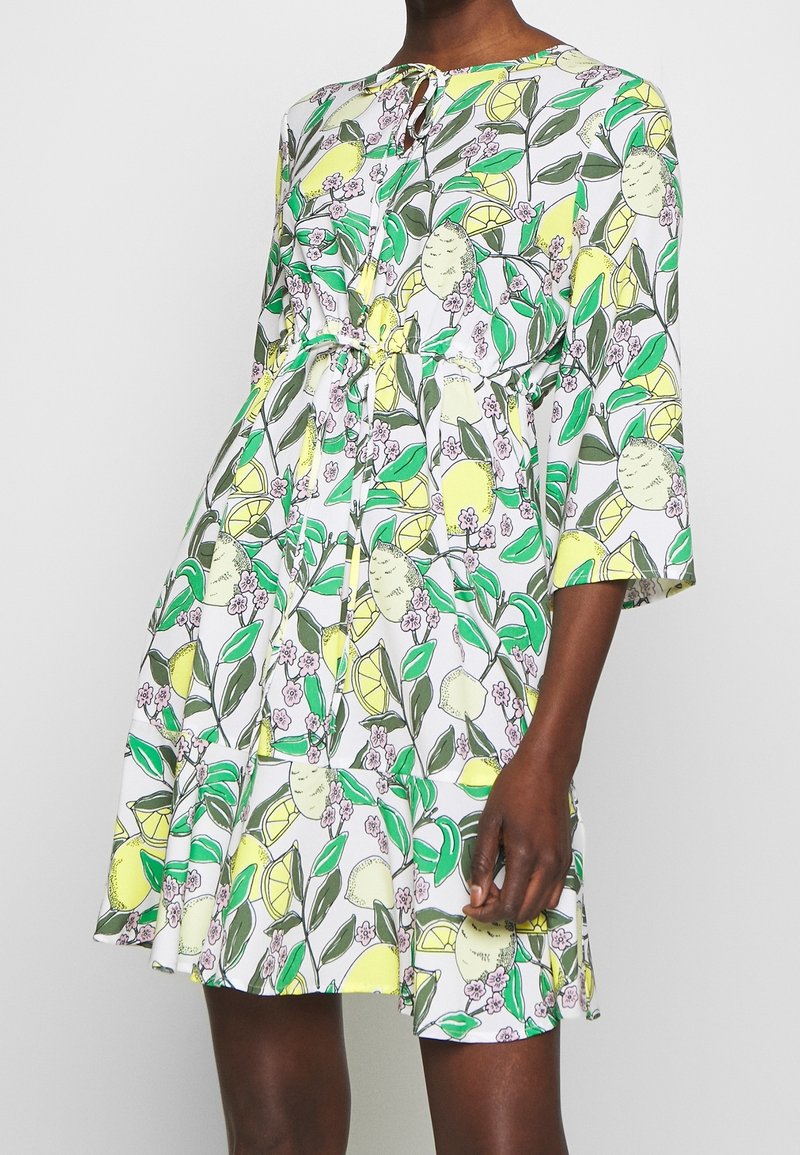 Robe blanche longueur genoux avec motif citron jaune et feuille verte, manches trois-quarts, encolure à nouer, et taille cintrée par une ceinture assortie.