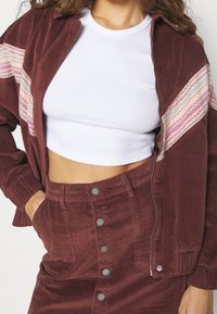 Vrouw in een witte crop top met een bordeaux corduroy jasje en een bijpassende knooprok met kleurrijke strepen op de mouwen van het jasje.