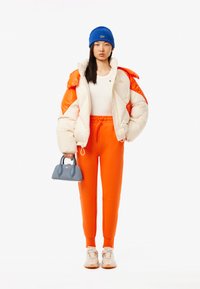 En pufferjacka i kräm och orange, vitt linne, orangea sweatpants, blå stickad mössa och en liten blå handväska. Sneakers med texturerade sulor.
