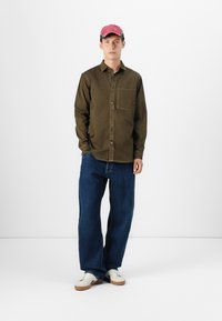 Camisa marrón de botones con un bolsillo en el pecho, combinada con pantalones vaqueros azules holgados y zapatillas blancas. La persona lleva una gorra rosa con acentos negros.