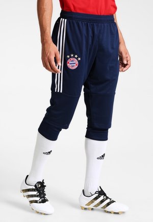 Homme portant un pantalon de sport bleu foncé avec des bandes blanches et le logo du FC Bayern Munich, des chaussettes blanches Adidas et des chaussures de football blanches Adidas.