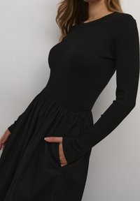 Czarna sukienka z dopasowanym, prążkowanym gorsetem i zwiewną, marszczoną spódnicą, z długimi rękawami i kieszeniami po bokach. Gładka faktura na całej powierzchni.