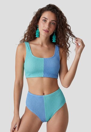 Frau mit lockigem Haar, die einen strukturierten, zweifarbigen blau-türkisfarbenen Bikini und passende Quastenohrringe trägt und ihr Haar berührt.