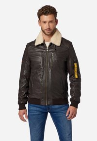 Braune Lederbomberjacke mit Schaffellkragen, Reißverschlusstaschen, gerippten Bündchen und Saum sowie einem gelben Akzent am linken Ärmel.